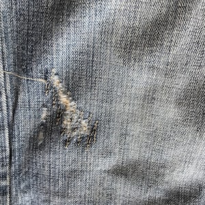 Peut inclure: Gros plan sur un tissu denim bleu avec une zone ab&icirc;m&eacute;e. Le tissu a un aspect us&eacute; avec une d&eacute;chirure, r&eacute;par&eacute;e avec des coutures noires. Le denim pr&eacute;sente un d&eacute;lavage classique.