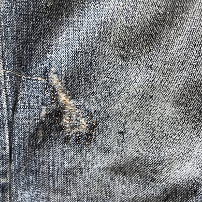 Peut inclure: Gros plan sur un tissu denim bleu avec une zone ab&icirc;m&eacute;e. Le tissu a un aspect us&eacute; avec une d&eacute;chirure, r&eacute;par&eacute;e avec des coutures noires. Le denim pr&eacute;sente un d&eacute;lavage classique.