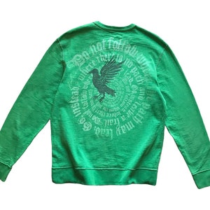 Puede incluir: Una sudadera verde con cuello redondo y mangas largas. La parte trasera presenta un diseño circular con un gráfico de cuervo y el texto "No sigas el camino. En cambio, ve donde no hay camino y deja un rastro."