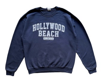 Puede incluir: Sudadera azul marino con cuello redondo y las palabras "HOLLYWOOD BEACH" en blanco en forma de arco. Debajo, un rectángulo más pequeño con la palabra "FLORIDA" en blanco. Estilo casual.