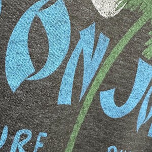 May include: Dark grey t-shirt featuring a blue and green tropical design. The design includes the words "ON J" in blue, a green palm tree, and the word "SHOP" in blue. A white moon is also visible.