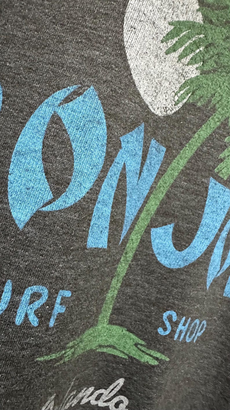 May include: Dark grey t-shirt featuring a blue and green tropical design. The design includes the words "ON J" in blue, a green palm tree, and the word "SHOP" in blue. A white moon is also visible.