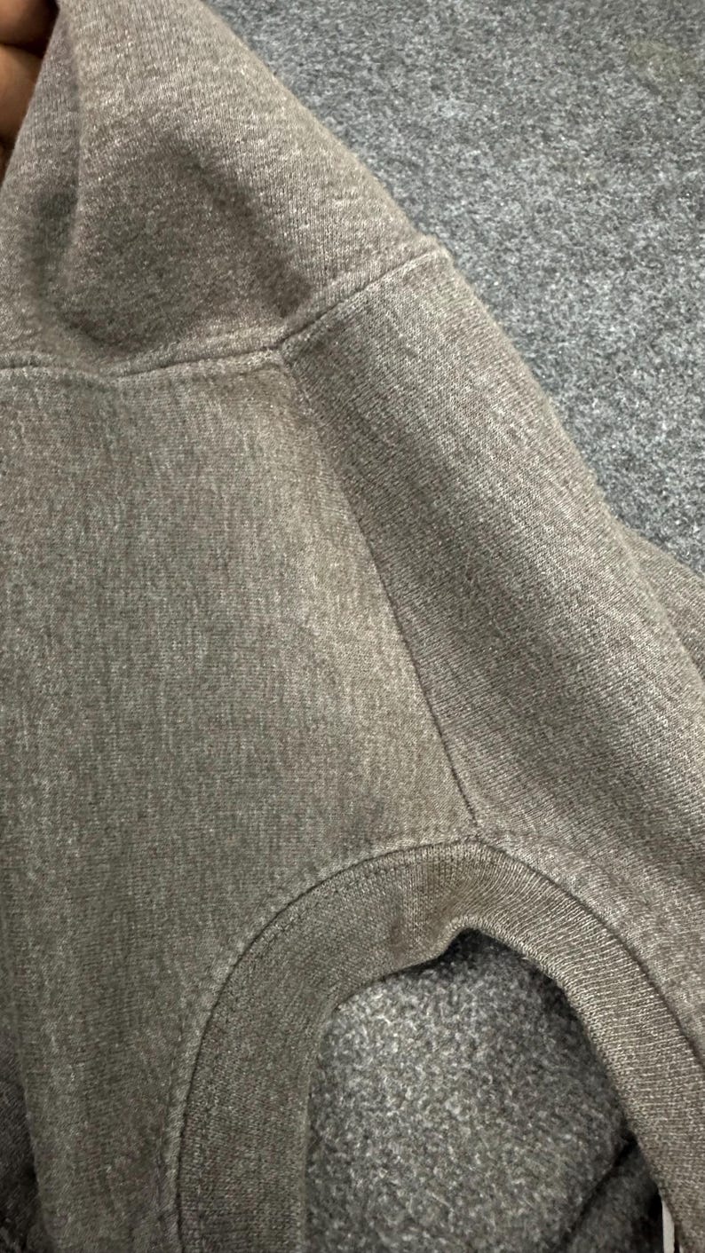 May include: Close-up of a grey hooded sweatshirt. The fabric is a soft, textured knit with visible stitching along the seams. The hood and shoulder area are in focus, showing the garment's construction.
