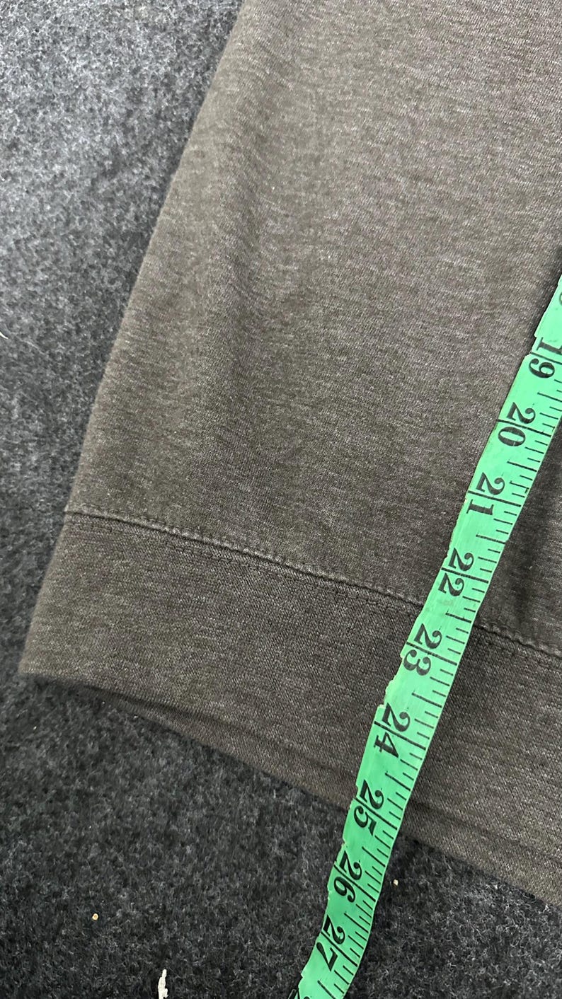 May include: Close-up of dark grey fabric with a sewn hem. A green measuring tape is laid across the fabric, showing measurements from 48 to 69 cm.