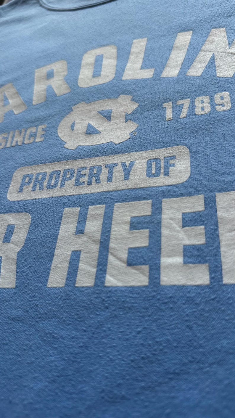 Peut inclure: Un t-shirt bleu clair avec du texte blanc qui dit "CAROLINA SINCE 1789 PROPERTY OF THE HEEL". Le t-shirt pr&eacute;sente un contour blanc du logo des Tar Heels de l'Universit&eacute; de Caroline du Nord.