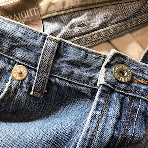 Peut inclure: Gros plan sur un jean en denim bleu avec des coutures visibles et des fermetures &agrave; boutons m&eacute;talliques. Le jean pr&eacute;sente un design classique avec un d&eacute;lavage, mettant en valeur les d&eacute;tails du tissu et de la construction. La marque Wrangler est visible sur les boutons.