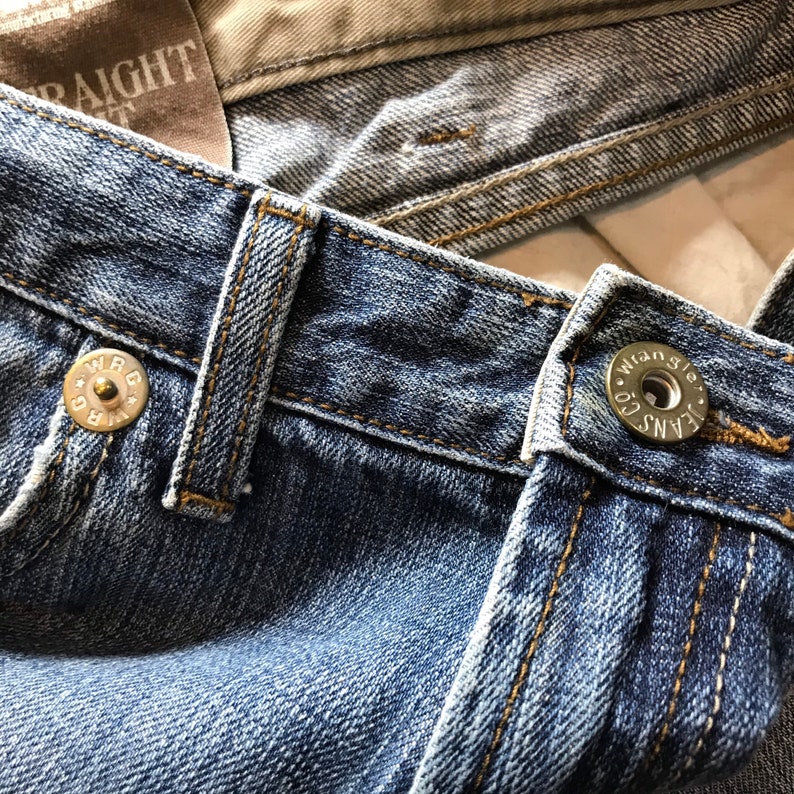 Peut inclure: Gros plan sur un jean en denim bleu avec des coutures visibles et des fermetures &agrave; boutons m&eacute;talliques. Le jean pr&eacute;sente un design classique avec un d&eacute;lavage, mettant en valeur les d&eacute;tails du tissu et de la construction. La marque Wrangler est visible sur les boutons.