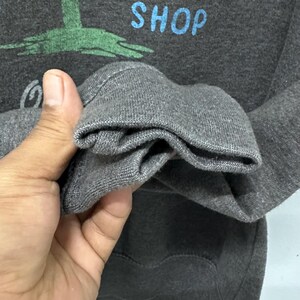 May include: Dark grey sweatshirt featuring the word "MOON" in blue, a green palm tree graphic, and the word "SHOP" in blue. The sleeve cuff is visible, showing the texture of the grey fabric.