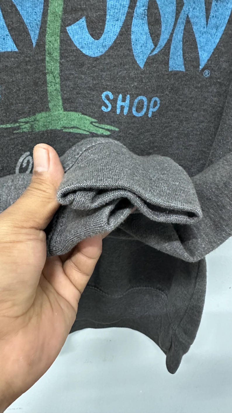 May include: Dark grey sweatshirt featuring the word "MOON" in blue, a green palm tree graphic, and the word "SHOP" in blue. The sleeve cuff is visible, showing the texture of the grey fabric.