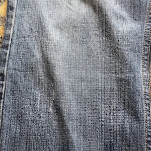 Peut inclure: Gros plan sur un tissu denim bleu avec une zone us&eacute;e. Le denim a un aspect d&eacute;lav&eacute; avec une texture visible et des coutures sur le bord. Le tissu montre des signes d'usure.