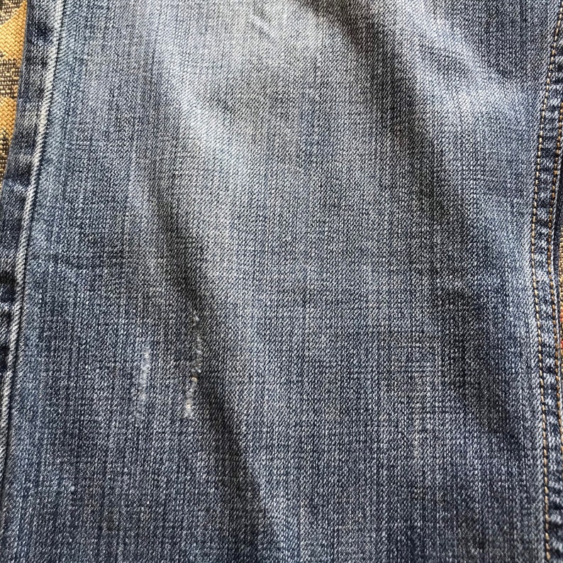 Peut inclure: Gros plan sur un tissu denim bleu avec une zone us&eacute;e. Le denim a un aspect d&eacute;lav&eacute; avec une texture visible et des coutures sur le bord. Le tissu montre des signes d'usure.