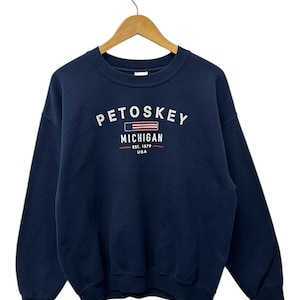 May include: Navy blue sweatshirt with the word "PETOSKEY" in white block letters above "MICHIGAN" and "EST. 1879 USA" in red and white. The sweatshirt is hanging on a wooden hanger.
