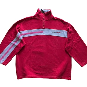 Peut inclure: Un sweat-shirt rouge Fubu Sport avec un col montant et une fermeture éclair quart de zip. Il présente une bande horizontale grise sur la poitrine et des rayures grises avec des accents roses sur les manches. Le logo Fubu Sport est imprimé sur la poitrine.