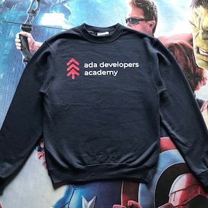 Op de afbeelding: Marineblauwe sweatshirt met de tekst "ada developers academy" in het wit en een rode afbeelding. De Champion-sweatshirt heeft lange mouwen.