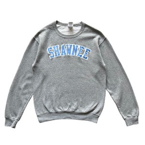 May include: A grey sweatshirt featuring the word "SHAWNEE" in light blue, arched across the chest with a white outline. The sweatshirt has a crew neck and long sleeves.