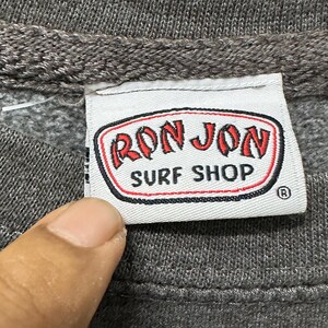 May include: Close-up of a grey sweatshirt label with the words "RON JON SURF SHOP" in red and white. The label has a black border and a registered trademark symbol. The sweatshirt is grey.