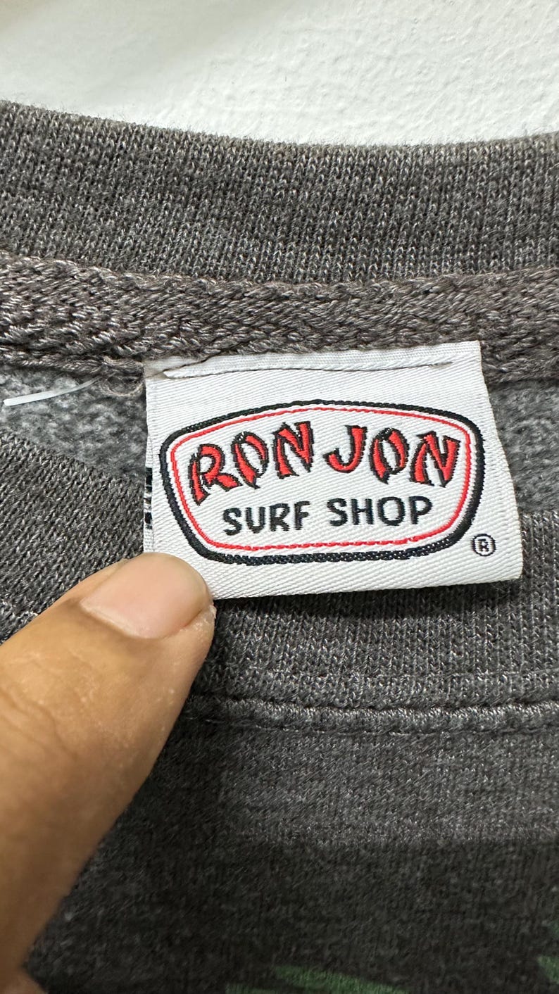 May include: Close-up of a grey sweatshirt label with the words "RON JON SURF SHOP" in red and white. The label has a black border and a registered trademark symbol. The sweatshirt is grey.