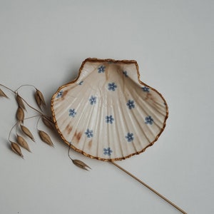 Könnte beinhalten: Eine weiße Muschel mit goldenem Rand und blauem Blumenmuster. Die Muschel ist eine dekorative Schale.