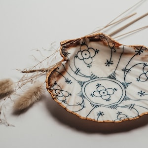 Op de afbeelding: Een witte schelpvormige schaal met een gouden rand en een blauw-wit bloemenpatroon. De schaal is versierd met een gestileerd bloemenpatroon.