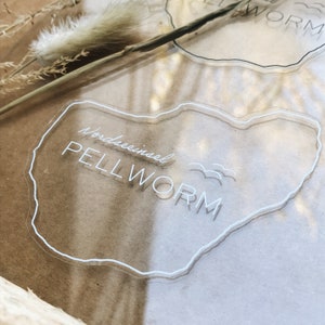 Könnte beinhalten: Ein transparenter Acrylaufkleber mit einer weißen Umrandung der Insel Pellworm, Deutschland. Der Text "Nordseeinsel PELLWORM" ist in weiß auf dem Aufkleber geschrieben.