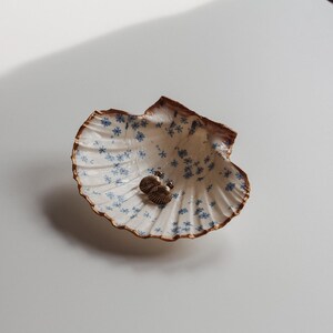 May include: A decorative seashell-shaped dish with a white interior adorned with blue floral patterns. The dish holds two small, gold-colored earrings. The shell has a brown, textured edge, and sits on a white surface.