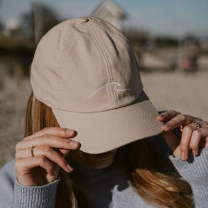 Può includere: Cappellino da baseball beige con visiera curva. Il cappellino presenta un motivo a onda bianca ricamata sul davanti. Realizzato in materiale morbido, ha un classico design a sei pannelli.