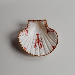 May include: A white scallop shell dish featuring a red lobster design. The shell has a gold-painted rim and a textured surface. The lobster design is repeated across the shell's surface.
