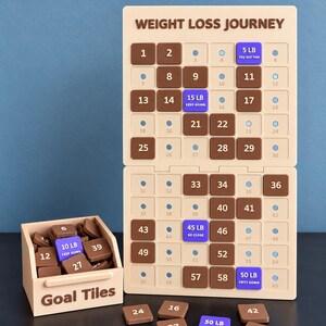 May include: A beige weight loss journey tracker board with brown numbered tiles and blue milestone tiles labeled 5 LB, 15 LB, 45 LB, and 50 LB. A matching beige box labeled Goal Tiles contains extra brown tiles, with several loose tiles scattered in front.