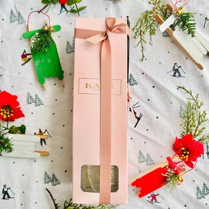 May include: A pink gift box tied with a matching ribbon, with the gold text "KAE". The box is surrounded by festive decorations, including miniature sleds, greenery, and red berries, all set against a winter-themed patterned background.