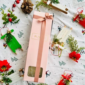 May include: A pink gift box tied with a ribbon, surrounded by festive decorations. The box features the letters "KA". Miniature sledges in red, white, and green, sprigs of greenery, red berries, and a pine cone are also present.