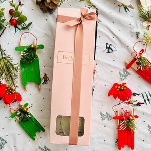 May include: A pink gift box with a gold ribbon and a clear window showing a glass diffuser bottle. The box is surrounded by small wooden sled ornaments decorated with pine sprigs and red berries. The box has the word "KATE" printed on the front.