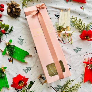 May include: A pink gift box with a gold ribbon and a clear glass bottle inside. The box is surrounded by various Christmas decorations, including red and white sleds, green pine branches, and red poinsettias.