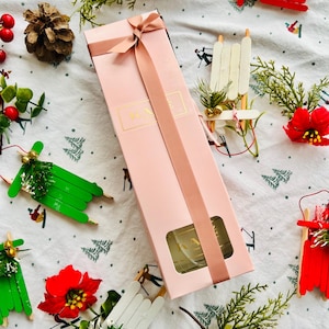 May include: A pink gift box with a gold ribbon and a clear glass bottle inside. The box is surrounded by various Christmas decorations, including red and white sleds, green pine branches, and red poinsettias.