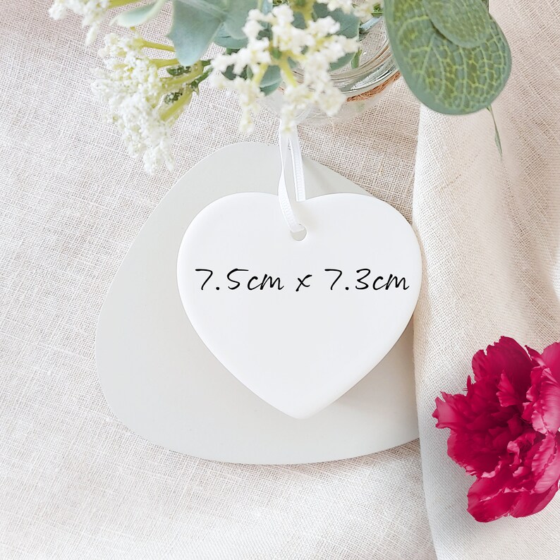 Puede incluir: Adorno de coraz&oacute;n de cer&aacute;mica blanca con una cinta blanca y el texto "7,5 cm x 7,3 cm" escrito en &eacute;l.