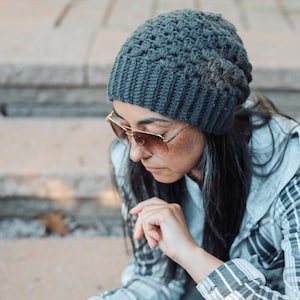 Puede incluir: Un gorro de ganchillo gris carbón oscuro con un patrón de puntos texturizados repetitivos en la corona y un borde acanalado. El gorro está confeccionado con un material grueso a base de hilo y se ajusta cómodamente a la cabeza.