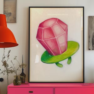 May include: A framed artwork featuring a large, pink, faceted gem atop a green, rounded base. The artwork is displayed on a white background, with a black frame. The image is set against a backdrop of a pink cabinet, an orange lamp, and other decorative items.