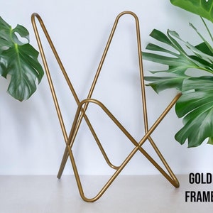 Op de afbeelding: Een gouden metalen stoelframe met een uniek ontwerp. Het frame is gemaakt van dun metaal en heeft een eenvoudige, moderne uitstraling. Het stoelframe staat op een wit oppervlak met een groene plant op de achtergrond. De tekst "GOLD FRAME" is zichtbaar in de rechterbenedenhoek van de afbeelding.