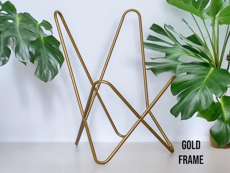 Op de afbeelding: Een gouden metalen stoelframe met een uniek ontwerp. Het frame is gemaakt van dun metaal en heeft een eenvoudige, moderne uitstraling. Het stoelframe staat op een wit oppervlak met een groene plant op de achtergrond. De tekst "GOLD FRAME" is zichtbaar in de rechterbenedenhoek van de afbeelding.