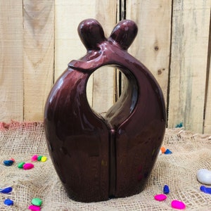 Può includere: Una scultura in ceramica marrone scuro di due figure che si abbracciano, formando un cuore con un ritaglio al centro.