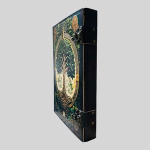 May include: A black leather-bound journal with a detailed illustration of a tree of life on the front cover. The cover features gold accents and intricate details. The journal has two metal clasps.