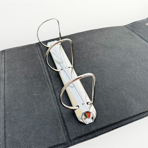 May include: A black three-ring binder with silver rings. The binder is closed and lying flat on a white surface.