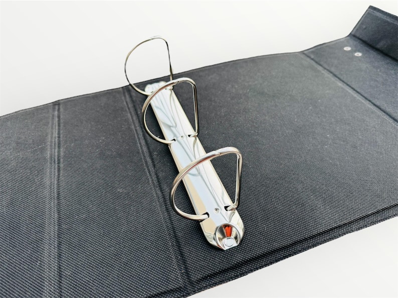 May include: A black three-ring binder with silver rings. The binder is closed and lying flat on a white surface.