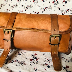 Puede incluir: Una bolsa de viaje de cuero marrón con dos correas ajustables y hebillas de latón. La bolsa está enrollada y tiene una costura alrededor del borde.
