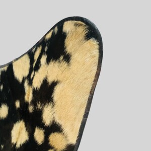 Op de afbeelding: Close-up van de rugleuning van een stoel van zwart en bruin koeienhuid. De stoel heeft een gebogen rugleuning en een zwarte leren rand.