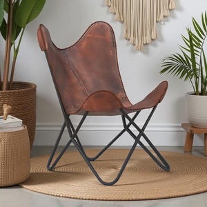 May include: A brown leather butterfly chair with a black metal frame. The chair is set on a round, woven jute rug. Potted plants and a woven side table with books complete the interior decor.
