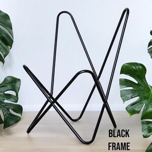 Op de afbeelding: Zwart metalen frame voor een stoel. Het frame is gemaakt van &eacute;&eacute;n stuk metaal dat gebogen en gelast is om de vorm van de stoel te vormen. Het frame is te zien op een houten vloer met groene planten op de achtergrond.