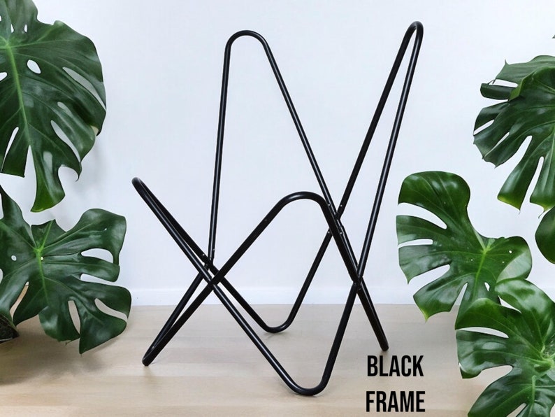 Op de afbeelding: Zwart metalen frame voor een stoel. Het frame is gemaakt van &eacute;&eacute;n stuk metaal dat gebogen en gelast is om de vorm van de stoel te vormen. Het frame is te zien op een houten vloer met groene planten op de achtergrond.