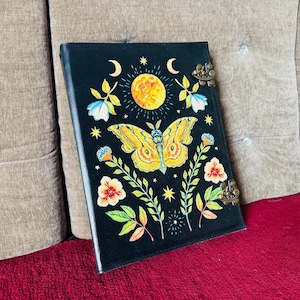 Puede incluir: Diario negro con un diseño de mariposa amarilla, una luna llena y detalles florales. El diario tiene un cierre de metal y está sobre una superficie roja.