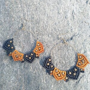 May include: A pair of hoop earrings with a black and orange macrame design. The earrings have a simple hoop shape and are decorated with a pattern of black and orange macrame knots. The earrings are a unique and stylish accessory.