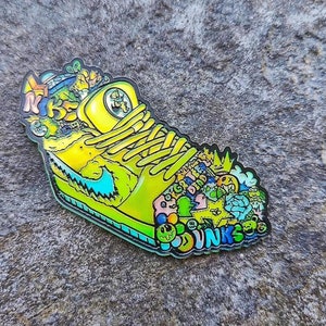 Könnte beinhalten: Eine gelbe und grüne Emaille-Anstecknadel mit einer Cartoon-Sneaker mit einer Vielzahl von bunten Kritzeleien und dem Wort "DUNKS" auf der Unterseite.